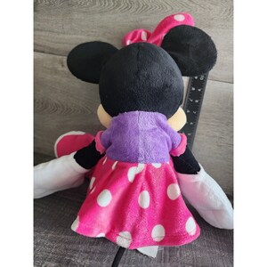 May include: A plush Minnie Mouse toy wearing a purple shirt and a pink polka dot dress. The toy has black ears and a red bow.