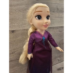 May include: A doll with long blonde hair wearing a purple dress with a sparkly design and a blue gem on the bodice.