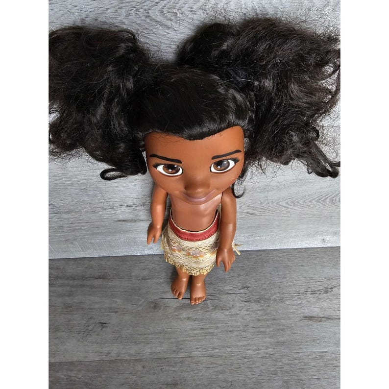 May include: A brown-skinned doll with long black hair styled in pigtails. The doll is wearing a brown and gold skirt and has a friendly expression.