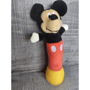May include: A plush Mickey Mouse toy with black ears, a tan face, and a red body. The toy has white gloves and a yellow patterned bottom. The toy is standing upright with its arms outstretched.