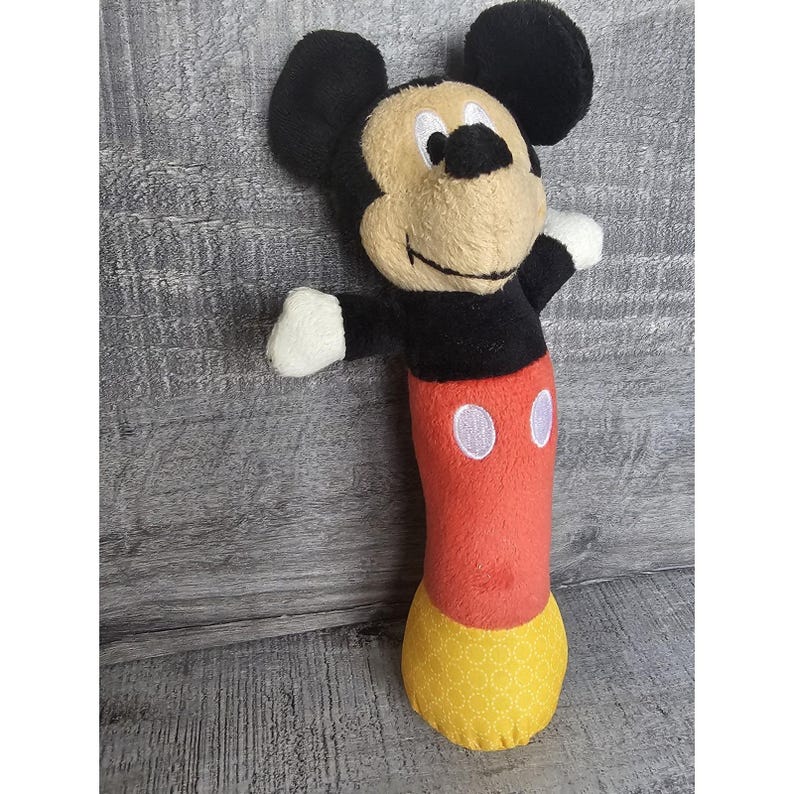 May include: A plush Mickey Mouse toy with black ears, a tan face, and a red body. The toy has white gloves and a yellow patterned bottom. The toy is standing upright with its arms outstretched.