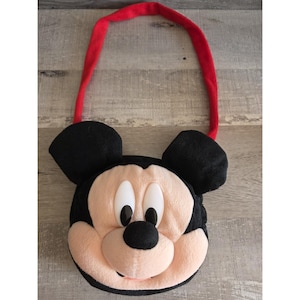 May include: A plush Mickey Mouse-shaped bag with black ears, a red strap, and a light pink face. The bag features Mickey's iconic black eyes, nose, and mouth. The bag is designed to be carried over the shoulder.