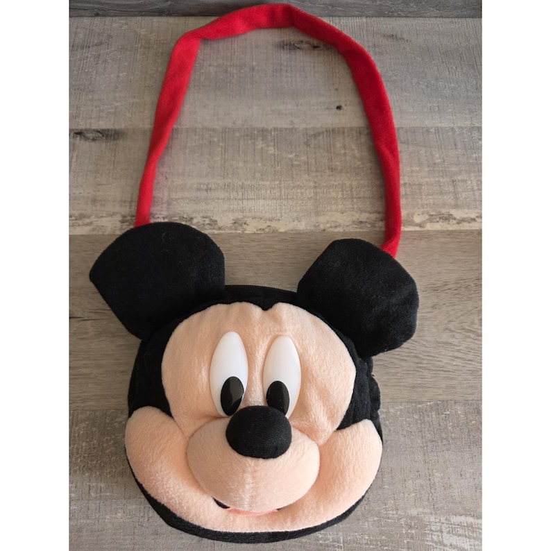 May include: A plush Mickey Mouse-shaped bag with black ears, a red strap, and a light pink face. The bag features Mickey's iconic black eyes, nose, and mouth. The bag is designed to be carried over the shoulder.