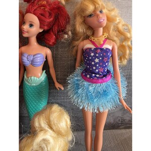 Op de afbeelding: Ariel-pop met rood haar, een paarse schelptop en een zeegroene zeemeerminstaart. Een blonde pop draagt een blauwe top met witte sterren, een roze rand en een blauwe franjerok. Een derde pop met blond haar is ook zichtbaar.
