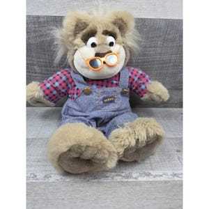 Puede incluir: Un león de peluche con gafas naranjas, una camisa a cuadros rosa y negro y un peto vaquero. El peto tiene un pequeño parche que dice "Guddi". El león tiene una melena marrón y grandes pies peludos.