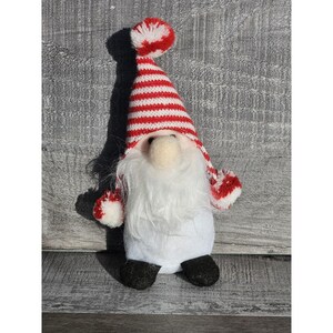 Puede incluir: Una figura de gnomo festiva con cuerpo blanco, larga barba blanca y pies negros. El gnomo lleva un gorro a rayas rojas y blancas con un pompón a juego y puños a rayas rojas y blancas. Es una decoración navideña.