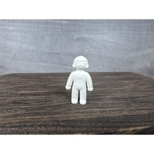 Könnte beinhalten: Kleine, weiße Kunststofffigur einer Person mit lächelndem Gesicht, kurzen Haaren und Knöpfen auf der Vorderseite. Die Figur steht auf einer dunkelbraunen Holzoberfläche vor grauem Hintergrund.