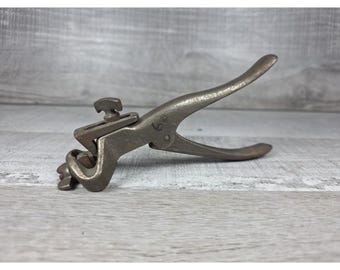 May include: A vintage metal saw set tool with a curved handle and adjustable mechanism. The tool is made of a dark, aged metal and is designed for adjusting saw blades. The overall design suggests a tool for woodworking or carpentry.