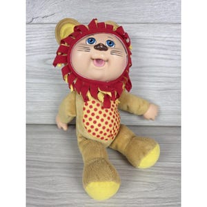 May include: A plush doll featuring a round face with blue eyes, a small nose, and a smiling mouth. The doll wears a red and yellow lion-themed hood with a mane, and a tan body suit with a red polka-dot pattern on the chest and yellow feet.