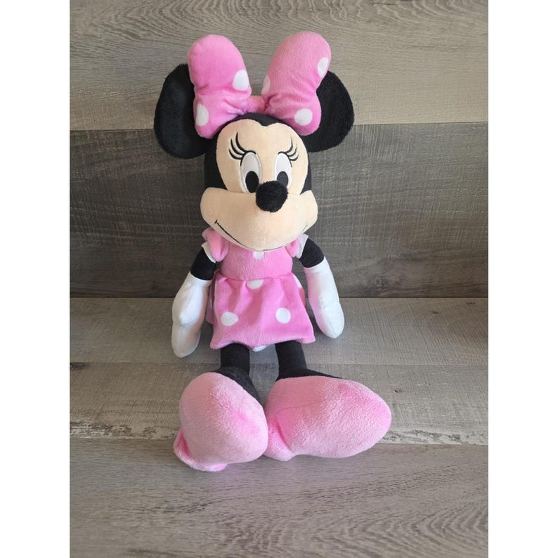May include: A plush Minnie Mouse doll. The doll has black ears, a pink bow with white polka dots, and a pink dress with white polka dots. The doll's shoes and hands are pink and white, respectively. The doll is approximately 18 inches tall.