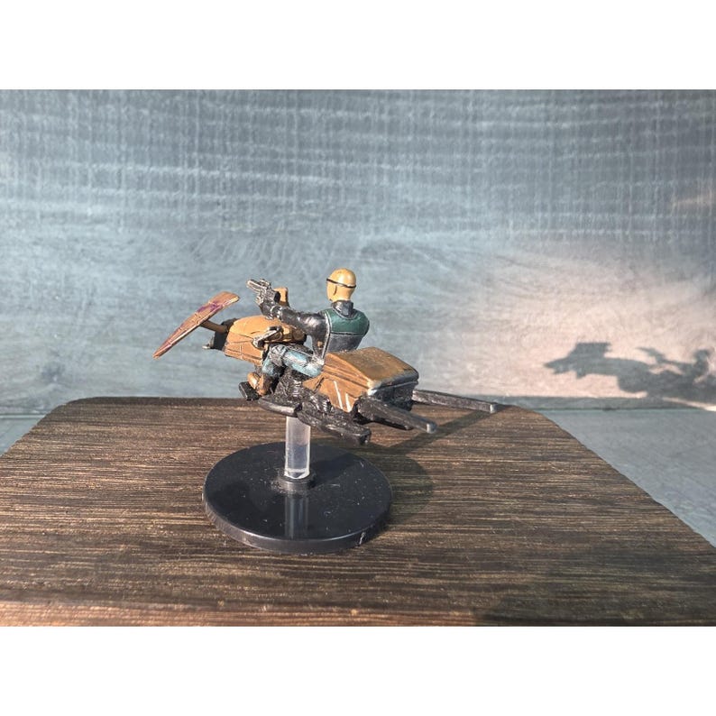 May include: A miniature figurine of a character riding a speeder bike. The figure is wearing a helmet and holding a blaster. The speeder bike is brown and black, with a clear stand. The background is a wooden surface.
