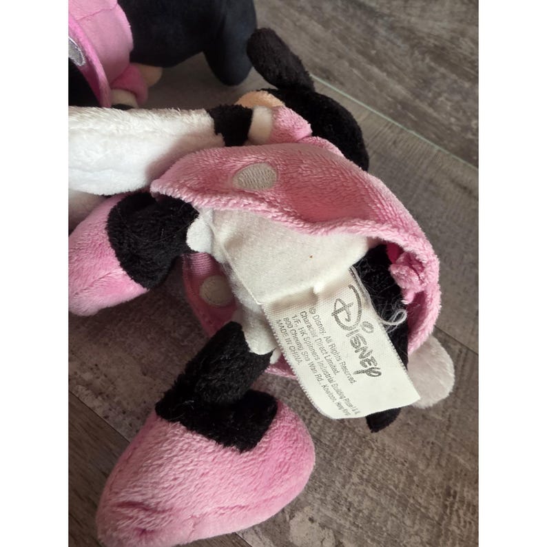 May include: A pink and black Minnie Mouse plush toy with a white face and a pink polka-dot bow. The toy has black ears and a pink dress. A Disney tag is attached.