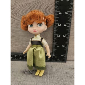 Puede incluir: Una mu&ntilde;eca peque&ntilde;a con cabello rojo, vestida con un vestido verde con un corpi&ntilde;o marr&oacute;n. La mu&ntilde;eca tiene ojos azules y una tez clara.