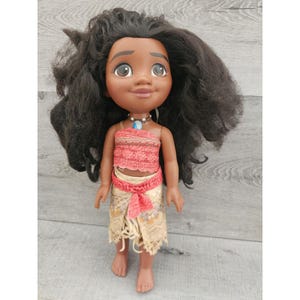 Puede incluir: Una mu&ntilde;eca de piel morena con el pelo largo y negro rizado que lleva un top rojo y blanco y una falda beige con un cintur&oacute;n marr&oacute;n. La mu&ntilde;eca lleva un collar azul y plateado.