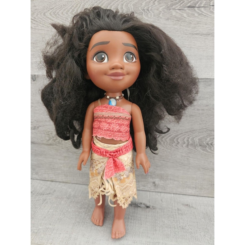 Puede incluir: Una mu&ntilde;eca de piel morena con el pelo largo y negro rizado que lleva un top rojo y blanco y una falda beige con un cintur&oacute;n marr&oacute;n. La mu&ntilde;eca lleva un collar azul y plateado.