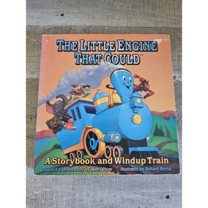 Könnte beinhalten: Ein Kinderbuch mit dem Titel "THE LITTLE ENGINE THAT COULD" mit einer Illustration eines blauen Zuges. Das Buchcover zeigt den Text "A Storybook and Windup Train" und ist nach der Geschichte von Watty Piper adaptiert, illustriert von Richard Bernal.