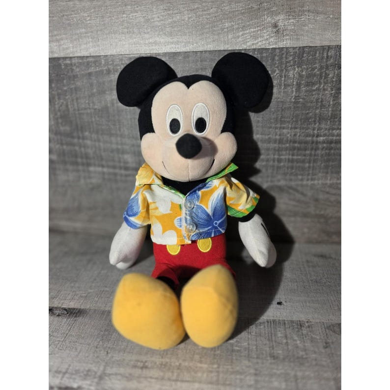 Puede incluir: Un peluche de Mickey Mouse con una camisa hawaiana con estampados florales amarillos, azules y verdes. El peluche tiene orejas negras, una cara blanca y pantalones cortos rojos. Se ven los zapatos amarillos del juguete.