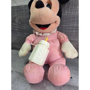 Puede incluir: Mu&ntilde;eca de peluche vintage de Minnie Mouse con un atuendo rosa con lunares y sosteniendo un biber&oacute;n blanco. La mu&ntilde;eca tiene orejas negras, una cara rosa y una boca roja. El biber&oacute;n tiene una tetina amarilla.