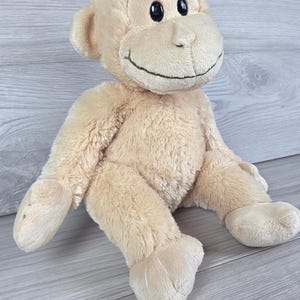 May include: A plush monkey toy made of soft, light beige fabric. The stuffed animal features a rounded head with small ears, black plastic eyes, and a stitched mouth forming a smile. It has long arms and legs with stitched details on the feet.