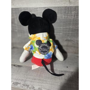 Puede incluir: Un peluche de Mickey Mouse, visto desde atr&aacute;s, con orejas y cabeza negras. El juguete lleva una colorida camisa hawaiana con un estampado floral y pantalones cortos rojos. Un parche en la camisa dice "7&ordm; Cumplea&ntilde;os de Jayden 15-07-2019."