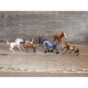 Puede incluir: Una colección de caballos de juguete en varios colores y poses. Los caballos incluyen blanco, marrón, gris y azul, algunos con marcas detalladas. Los juguetes están dispuestos sobre una superficie de madera, mostrando sus diferentes posturas.