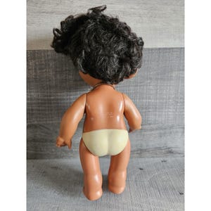 Pu&ograve; includere: Una bambola dalla pelle marrone con capelli neri ricci che indossa un pannolino giallo. La bambola &egrave; in piedi su una superficie di legno.