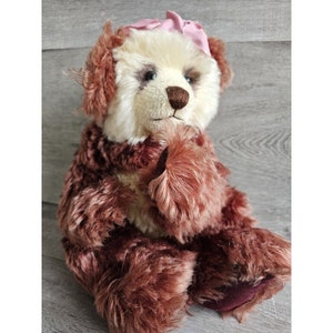 Può includere: Un orsacchiotto marrone e bianco con un fiocco rosa seduto su una superficie di legno. L'orso ha un naso marrone e occhi neri.