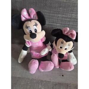May include: Two Minnie Mouse plush toys. The plush toys have black ears, black hair, and pink bows with white polka dots. They are wearing pink dresses and shoes. One is sitting upright, and the other is sitting down.