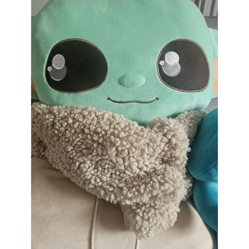 May include: A plush toy of a green character with large eyes and a brown fur scarf. The character is smiling and has a light green body.