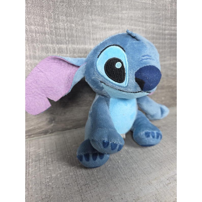 May include: A plush toy character featuring a blue body with a lighter blue chest patch, large ears with light purple inner sections, a dark blue nose, and embroidered black eyes. The character has a seated posture with visible paw pads.