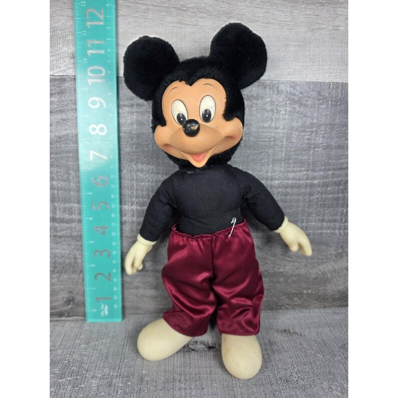May include: Vintage Mickey Mouse plush doll with black fur ears and head, a black shirt, and burgundy satin pants. The doll has cream-colored hands and feet. A ruler is next to the doll, showing measurements in inches.