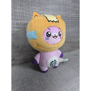 May include: A plush toy featuring a purple character wearing an orange hood with cat-like ears. The character has a white circular patch on its chest with a large letter F inside. It has embroidered facial features, including wide eyes and a small mouth.