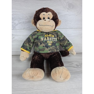 May include: A plush monkey toy with dark brown fur and a tan face, hands, and feet. It wears a green, brown, and black camouflage-patterned shirt with yellow trim. The shirt features the text ALL-STAR VARSITY TEAM 1997 printed in yellow letters.