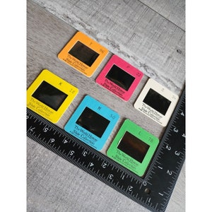 May include: A selection of colourful vintage slide film holders. The holders are in orange, pink, yellow, blue, green, and white. Each has a black window and the text "The World History Slide Collection". Numbers and letters are printed on each holder.