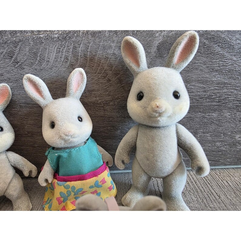 Puede incluir: Una colecci&oacute;n de figuras de conejos grises con orejas forradas de rosa. Un conejo lleva una camiseta turquesa y una falda amarilla con estampado floral. Los conejos tienen ojos negros y est&aacute;n de pie. El fondo es una superficie de madera gris.