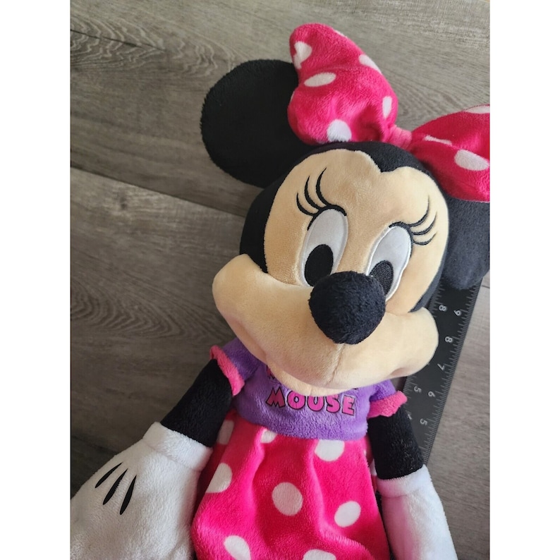 May include: A plush Minnie Mouse toy wearing a pink dress with white polka dots and a purple shirt with the text "MOUSE". She has a pink bow with white polka dots on her head.