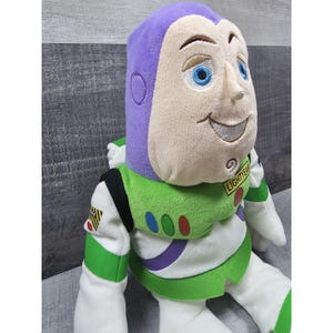 May include: A soft toy of Buzz Lightyear, with a purple helmet, a smiling face with blue eyes, and a green and white spacesuit. The doll has the word "LIGHTYEAR" on the chest and is made of soft, plush material.