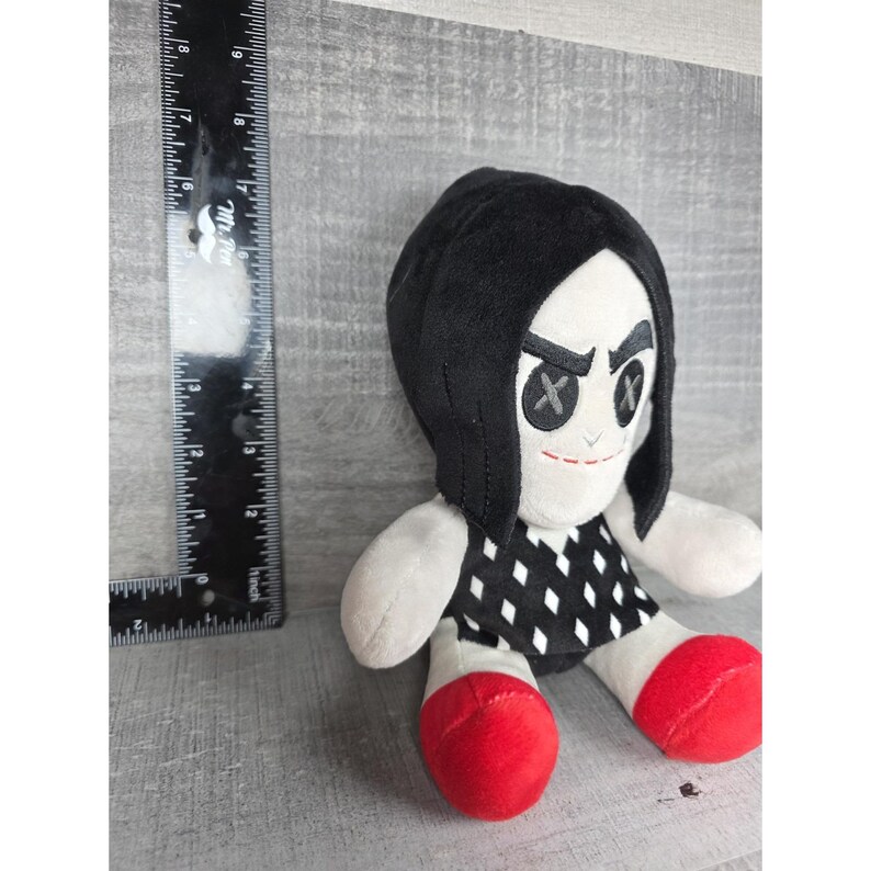 May include: A plush doll with a black head covering and a stern expression. The doll has a black and white diamond pattern dress, white arms, and red feet. A ruler is next to the doll.