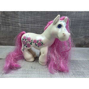May include: A white toy pony with pink hair and a pink rose design on its side. The pony has blue eyes and a light pink nose. The pony's hair is long and flowing, with a glittery sheen. The toy is standing on a wooden surface.
