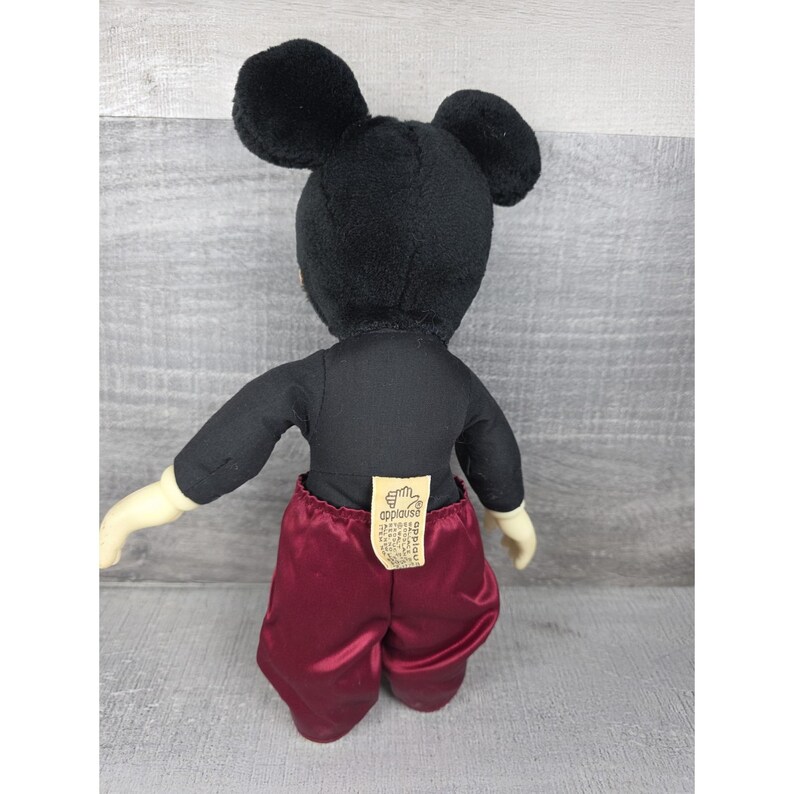 May include: A vintage Mickey Mouse plush toy, viewed from the back. The plush features a black head with large ears, a black shirt, burgundy satin shorts, and cream-colored hands. A tag is attached to the shorts.