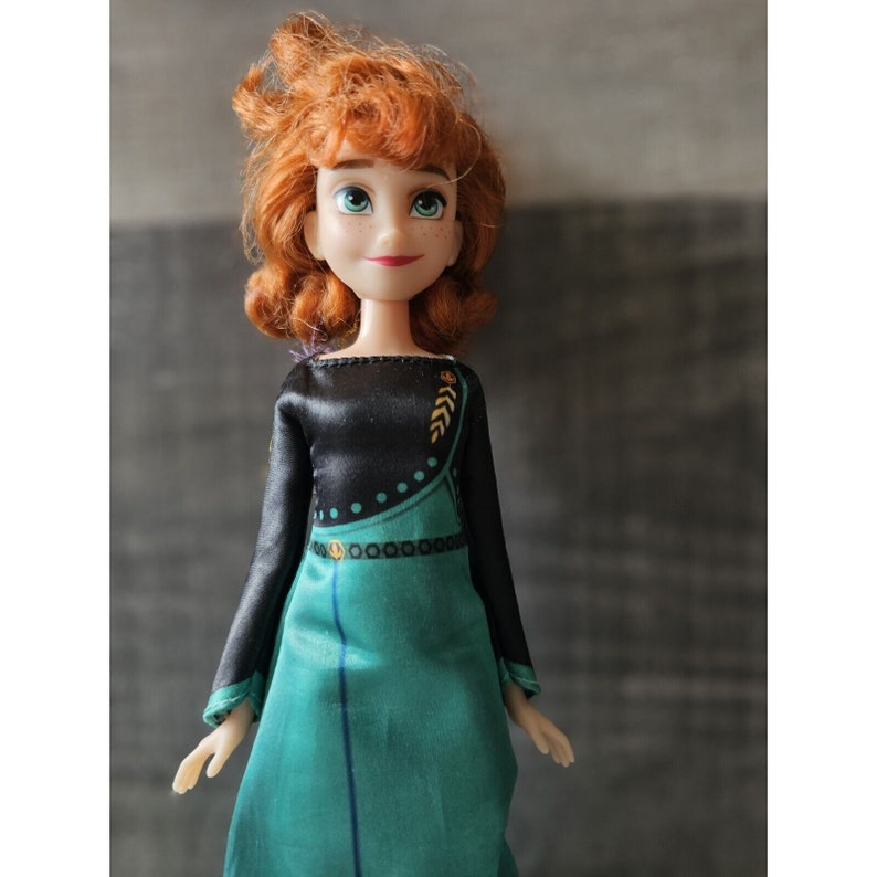 May include: A red-haired doll wearing a black and green dress with gold accents. The doll has a detailed face with blue eyes and a friendly expression.