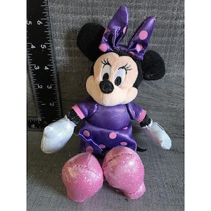 Op de afbeelding: Een Minnie Mouse knuffel. De knuffel heeft een paarse jurk met roze stippen, een bijpassende strik en roze glitter schoenen. Het heeft zwarte oren, witte handschoenen en een lachend gezicht. De knuffel is ongeveer 10 cm hoog.