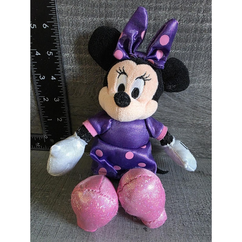 Op de afbeelding: Een Minnie Mouse knuffel. De knuffel heeft een paarse jurk met roze stippen, een bijpassende strik en roze glitter schoenen. Het heeft zwarte oren, witte handschoenen en een lachend gezicht. De knuffel is ongeveer 10 cm hoog.