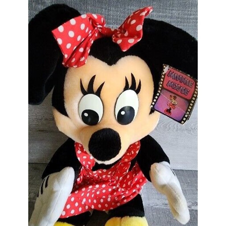 Pu&ograve; includere: Un pupazzo di peluche di Minnie Mouse che indossa un vestito rosso a pois bianchi e un fiocco rosso a pois bianchi. Il pupazzo ha occhi neri, naso nero e bocca nera. Il pupazzo tiene un cartello che dice "Minnie Mouse Movie".