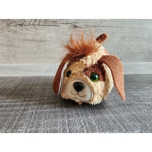 May include: A soft dog toy in shades of tan and brown. The toy features large, drooping ears, green eyes, and a tuft of brown hair. The dog's face is embroidered with a black nose and a smiling mouth.