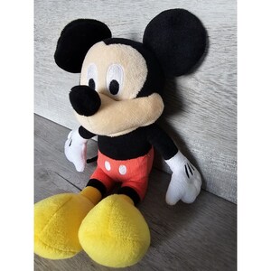 Puede incluir: Un juguete de peluche de Mickey Mouse con orejas negras, nariz negra y zapatos amarillos. El juguete lleva pantalones cortos rojos con botones blancos.