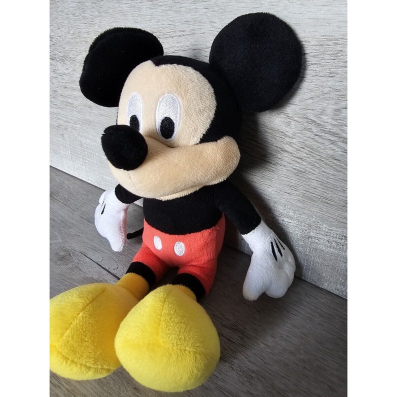 Puede incluir: Un juguete de peluche de Mickey Mouse con orejas negras, nariz negra y zapatos amarillos. El juguete lleva pantalones cortos rojos con botones blancos.
