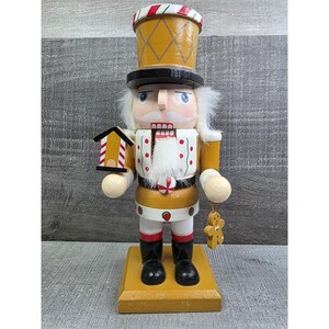 May include: A wooden Christmas nutcracker figurine. The figurine is painted in gold, white, black, and red. It has a white beard and hair, a black top hat, and holds a small house and a gingerbread man. The nutcracker stands on a gold base.
