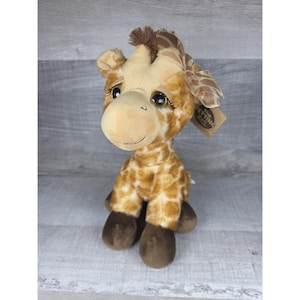 May include: A plush giraffe toy with a tan and brown spotted pattern. The giraffe has a light brown face, dark brown hooves, and a brown bow. The toy has large, expressive eyes and a tag that reads "The Getting Zoo".
