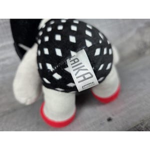 May include: A soft toy featuring a black and white diamond pattern on its back. The toy has white limbs and red soles. A tag with the word "LAIKA" is attached to the toy.